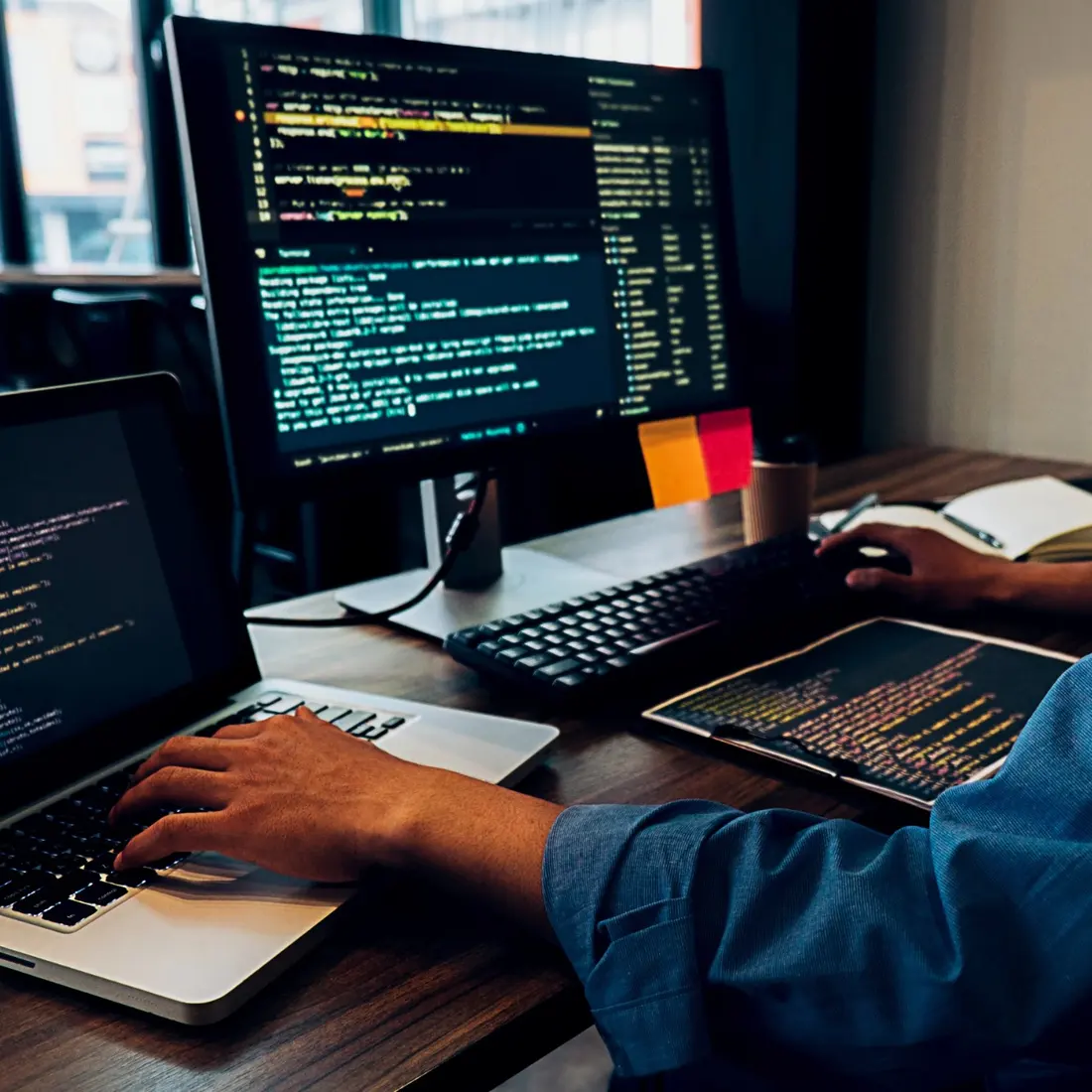 A web developer writing code at a laptop and monitor